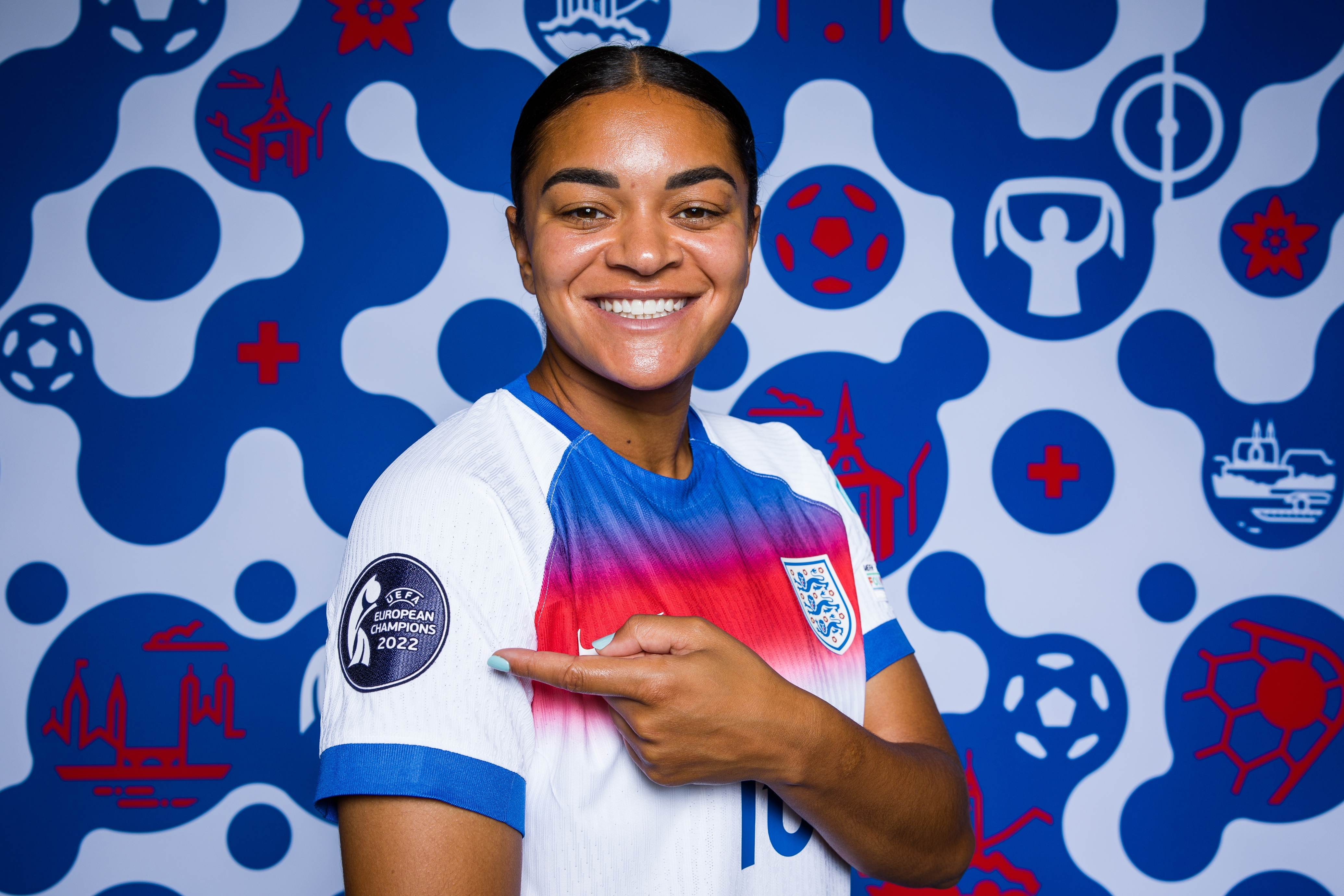 Person in Team England soccer jersey points to a Euro 2022 patch on their sleeve, smiling in front of a sports-themed backdrop