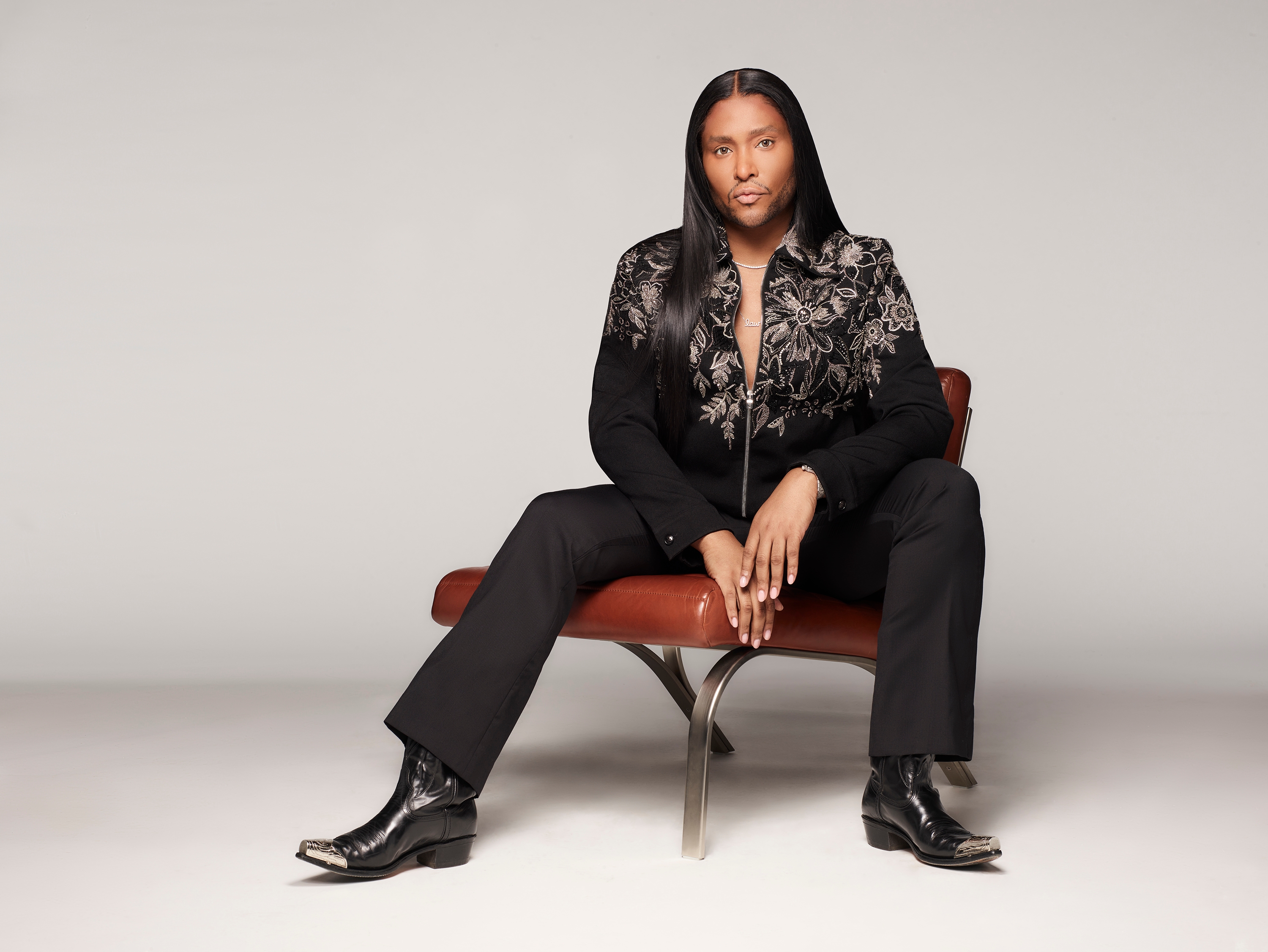 Person seated, wearing a black floral-patterned shirt, black pants, and boots, in a confident pose against a neutral background