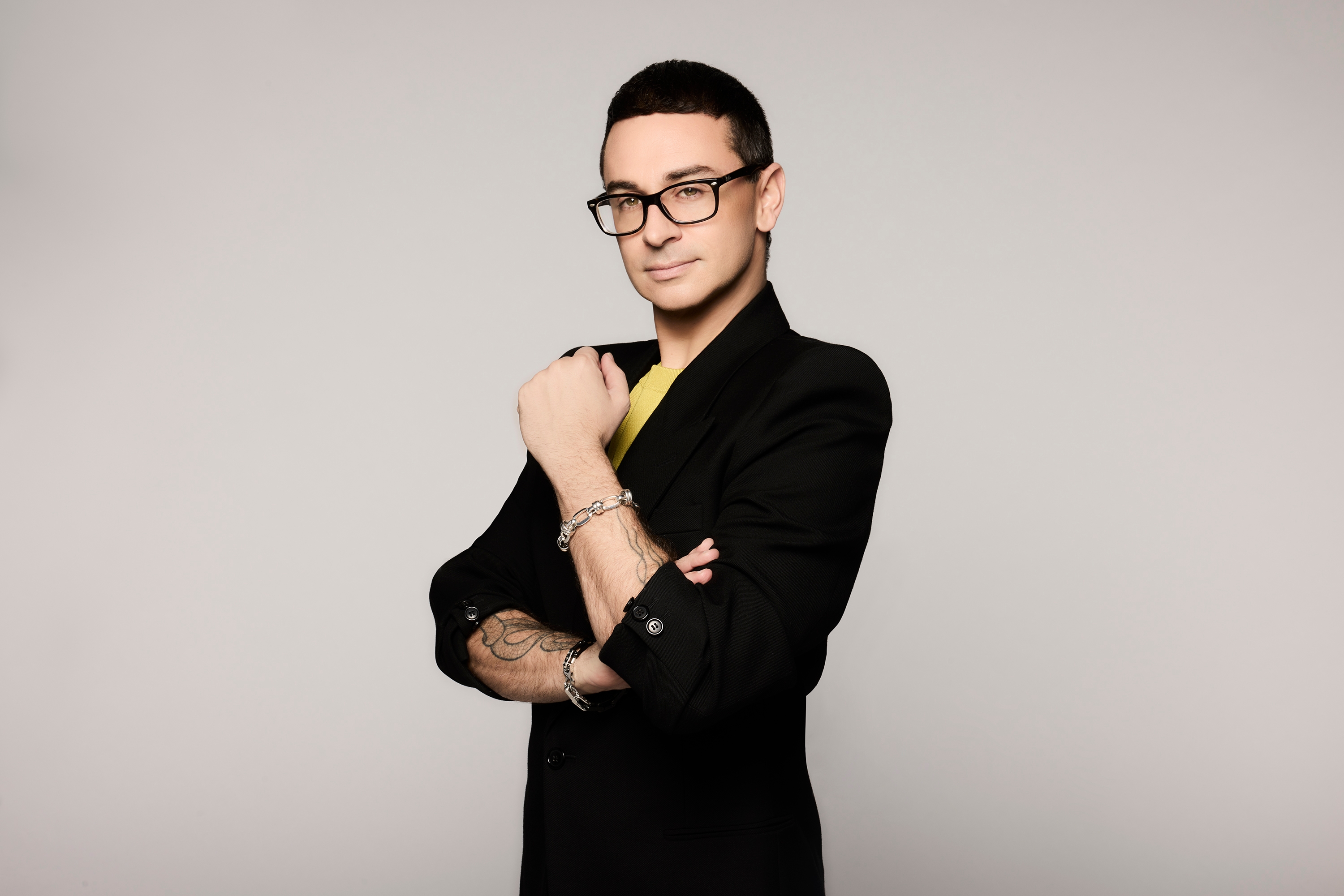 Person confidently posing in stylish glasses and a tailored blazer, displaying tattoos and bracelets, with a neutral background