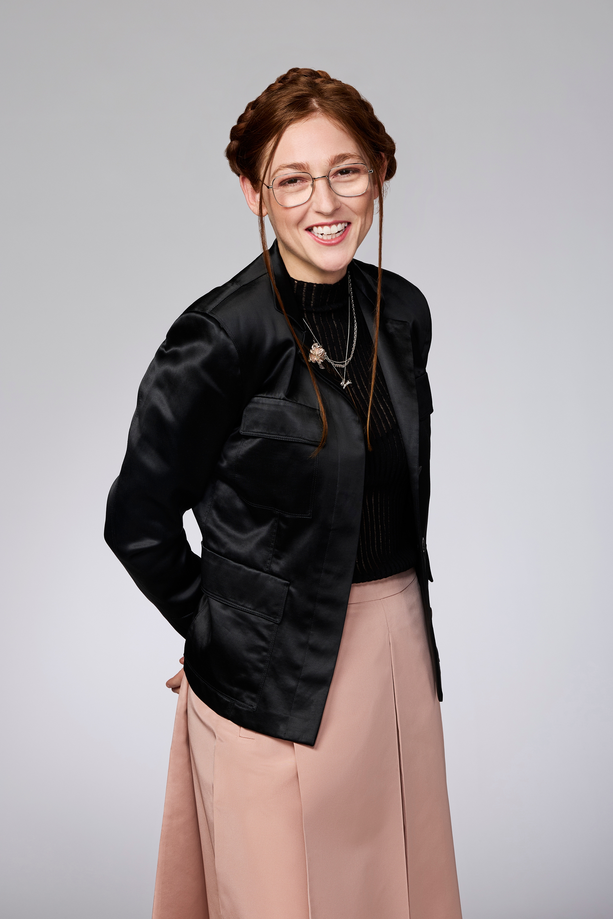 Person in a black jacket, pink skirt, and glasses, standing and smiling, hands behind their back, against a plain background
