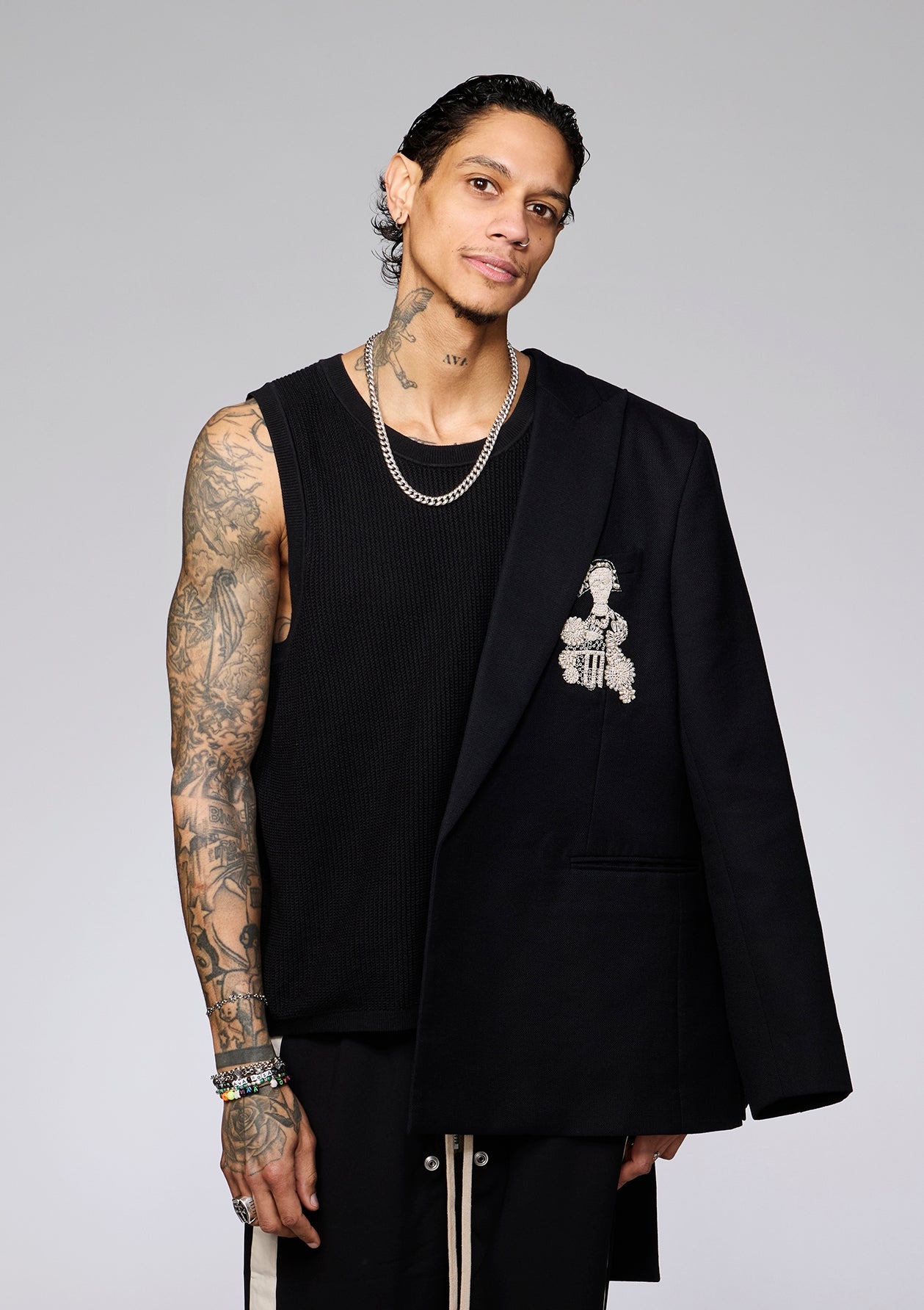 Person in casual-stylish outfit with sleeveless top, track pants, and blazer draped over shoulder, standing on a plain background
