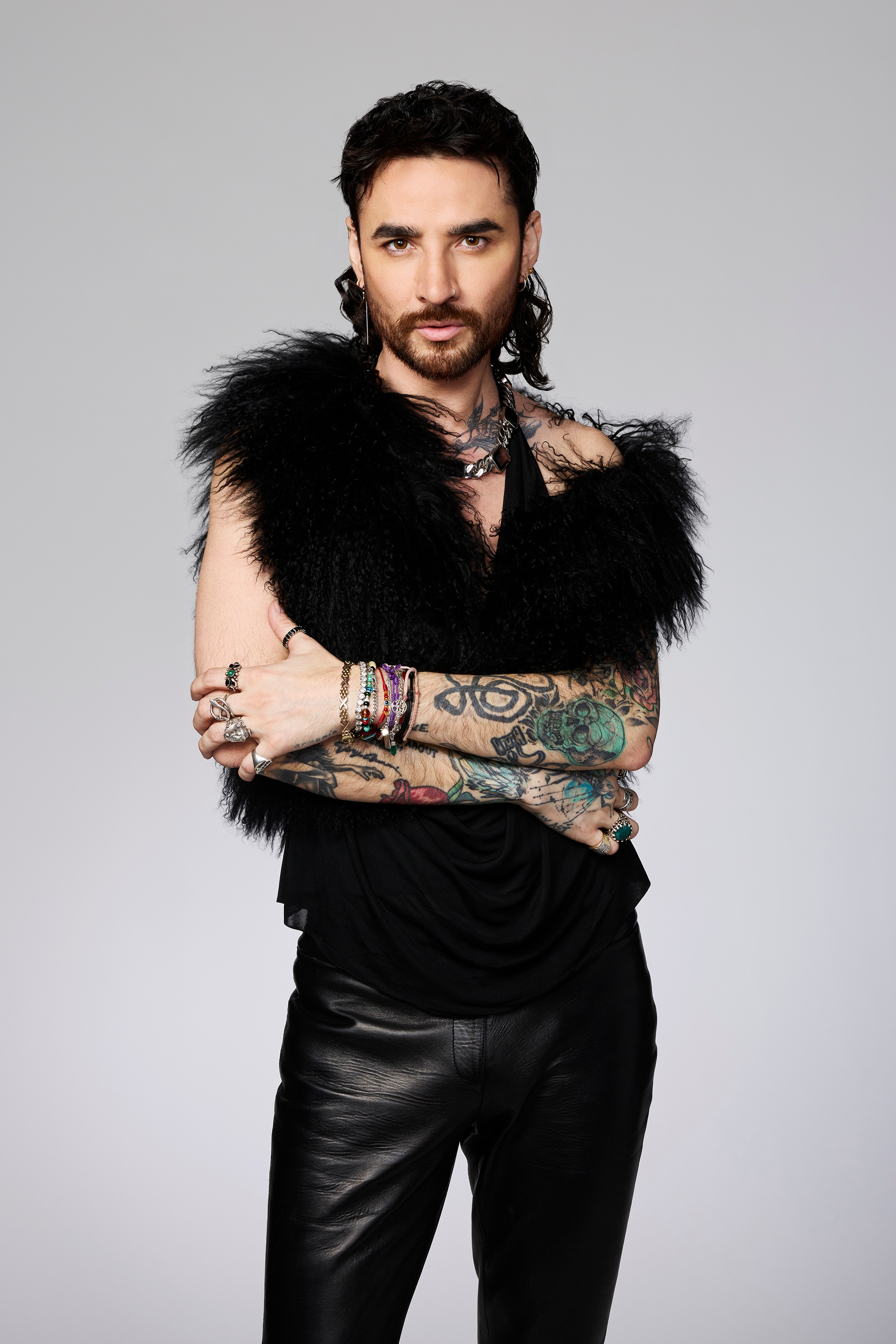 Person in a feathered top and leather pants, arms crossed, showcasing tattoos and jewelry; modern, edgy fashion style