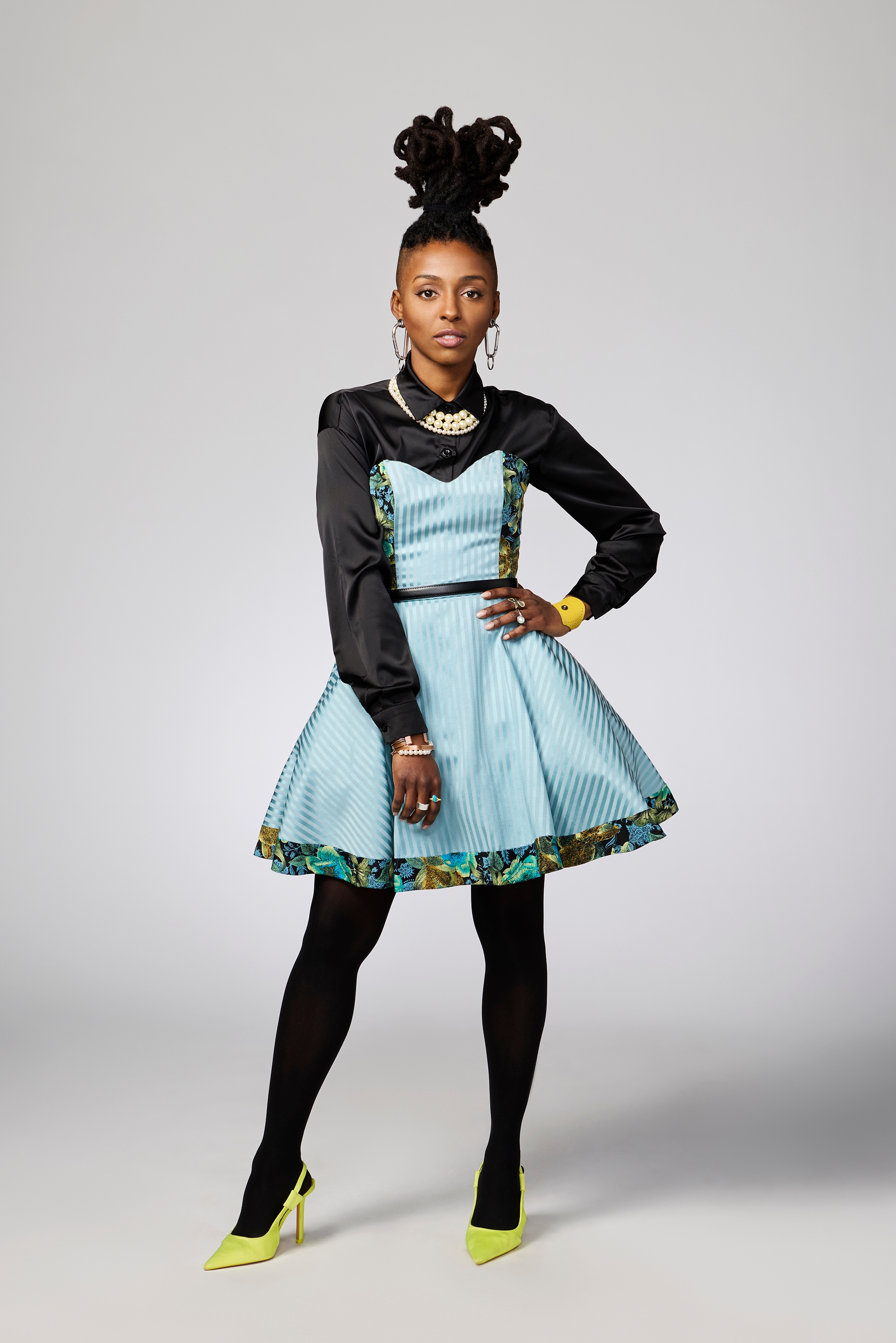 A person poses confidently, wearing a blue patterned dress with black accents, black tights, and neon heels, accessorized with jewelry