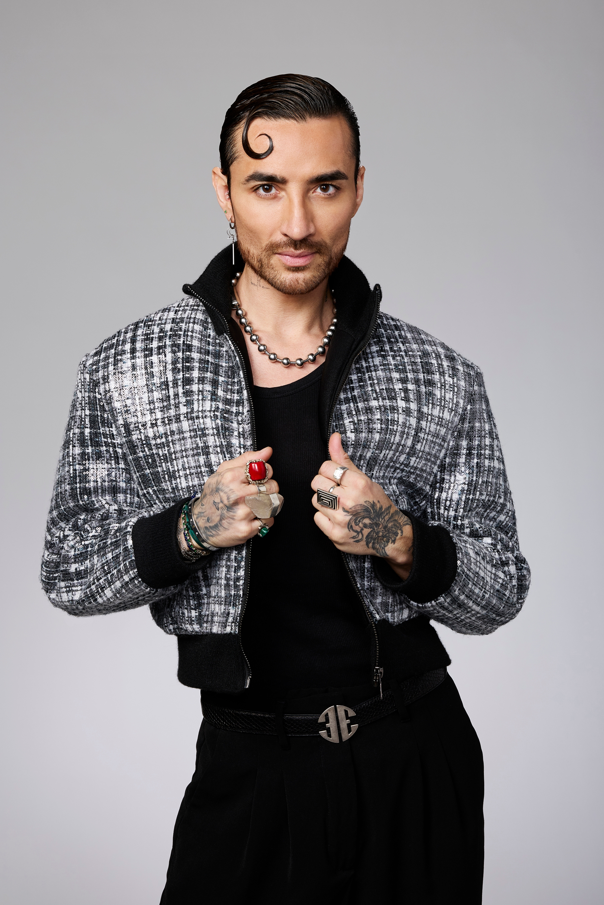 Person in a stylish checkered jacket, black shirt, and pants, with tattoos and jewelry, posing confidently