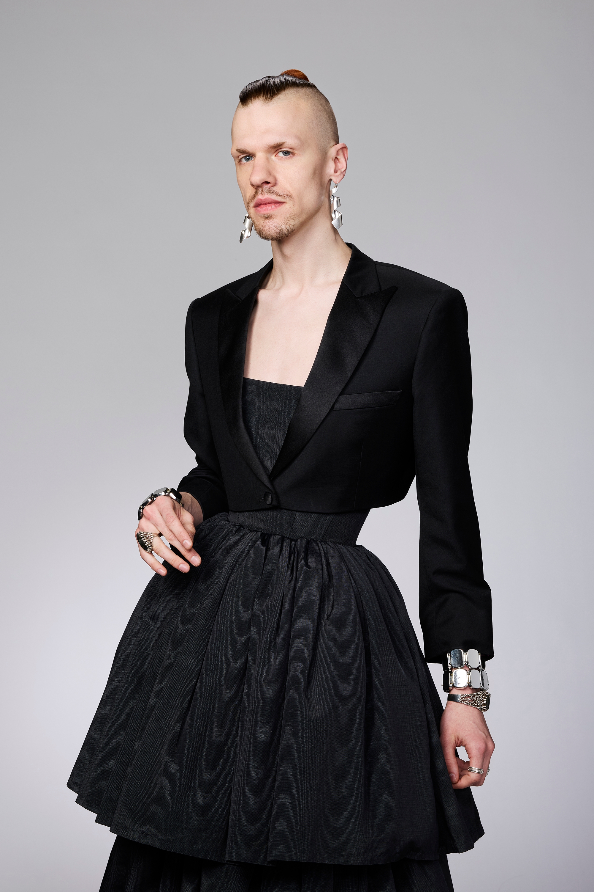Person wearing a structured black blazer and a patterned black skirt, with bold silver accessories, posing against a plain backdrop