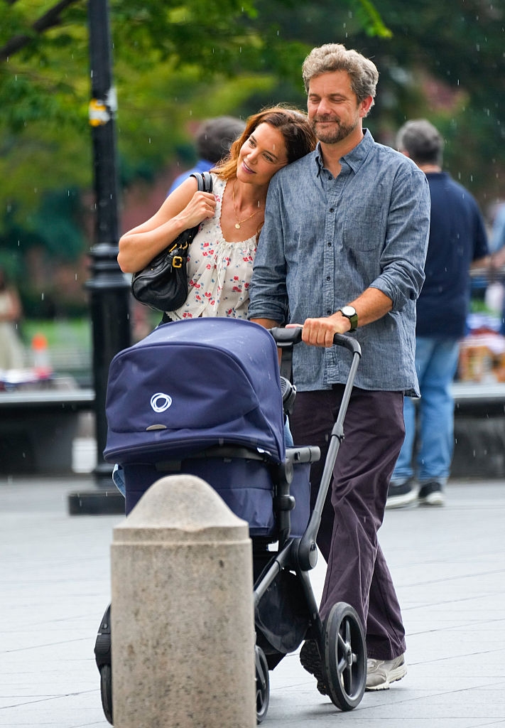 A couple strolls with a baby stroller outdoors. The woman leans affectionately on the man's shoulder, both appearing content and relaxed