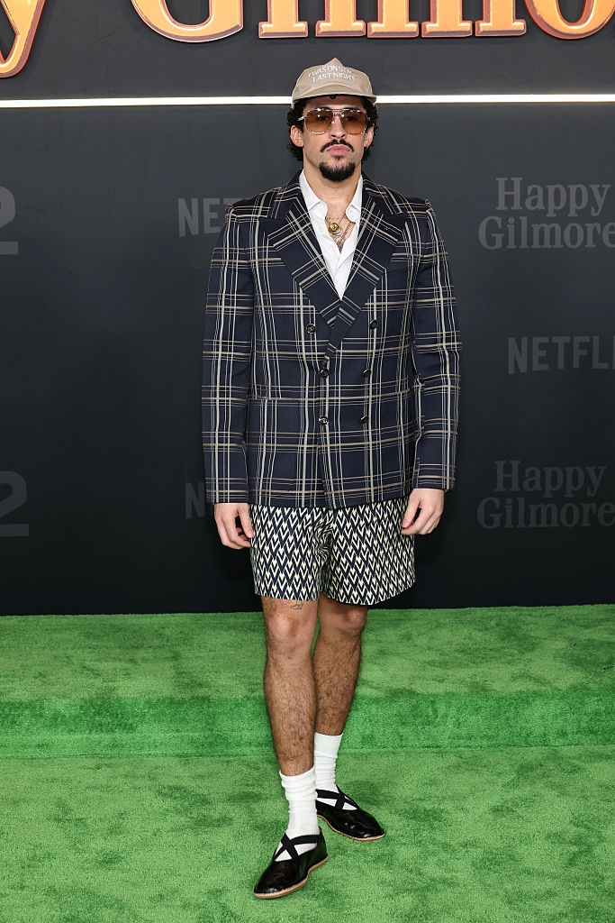 Person in plaid suit jacket, patterned shorts, and hat on a green carpet at a Netflix event