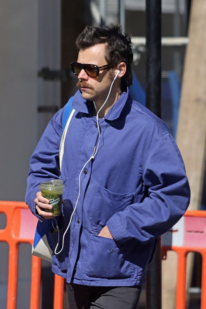 Person in a casual blue jacket, wearing sunglasses and earphones, holds a green drink while walking outdoors