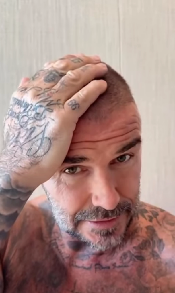 A heavily tattooed man with a shaved head and beard looks up while touching his head