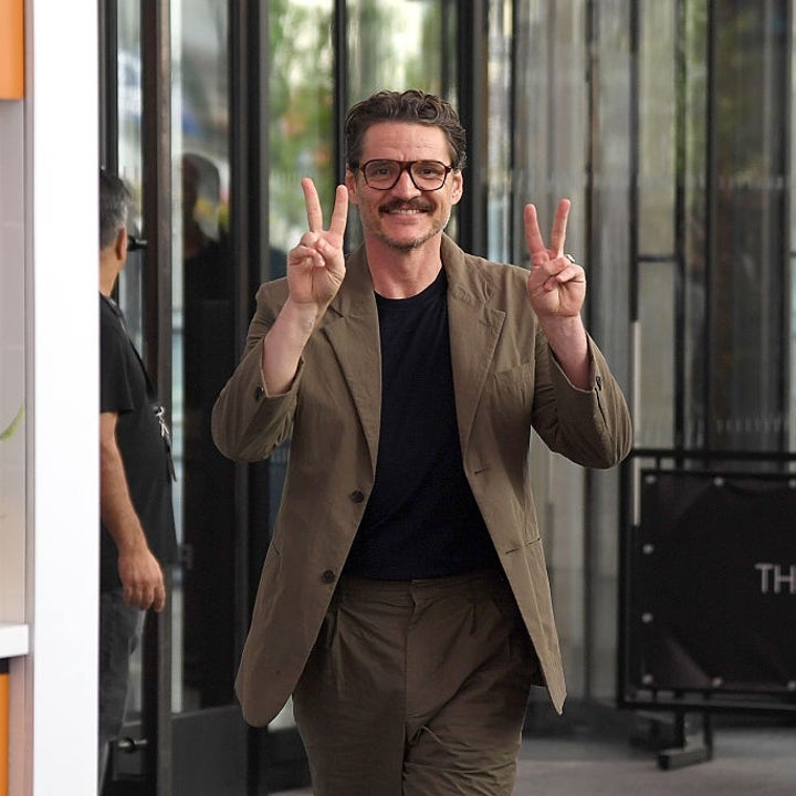 Person smiling, wearing a casual suit, giving peace signs with both hands, walking past a plant in a yellow pot