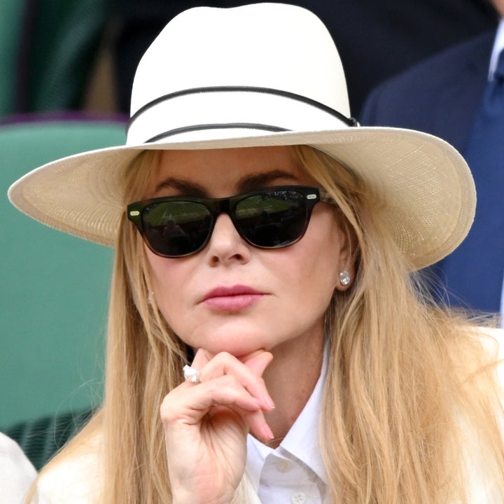 Nicole Kidman wearing a wide-brimmed hat, sunglasses, and a suit, sitting thoughtfully with a hand on their chin at an outdoor event