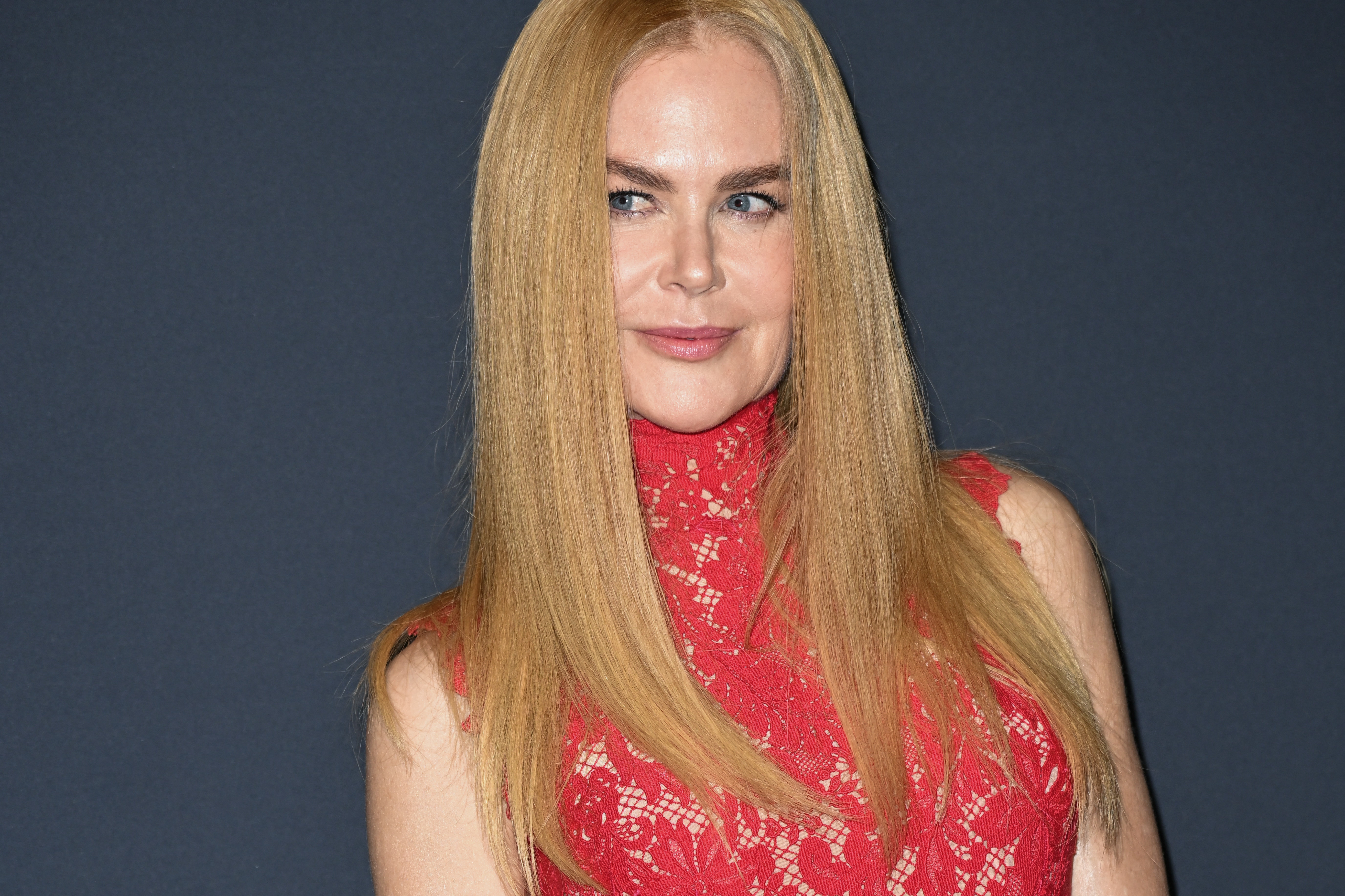 Nicole Kidman in a high-neck lace dress, gazing to the side with sleek, straight hair, on a media event backdrop