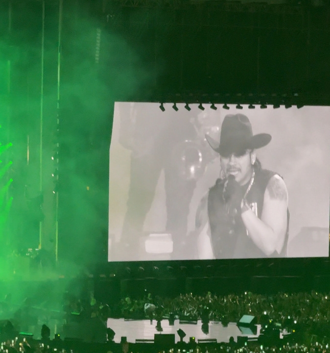 Cantante en concierto, usando un sombrero vaquero y chaleco, proyectado en una gran pantalla ante un público entusiasta