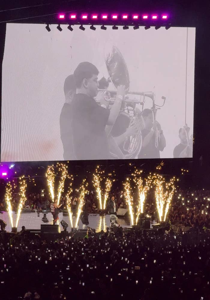 Concierto en vivo con fuegos artificiales; banda musical tocando frente a gran pantalla con imagen de trompetista. Público capturando el momento