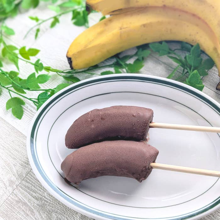 皿にチョコレートで覆われた2本のバナナがあり、背景に数本のバナナが置かれている。