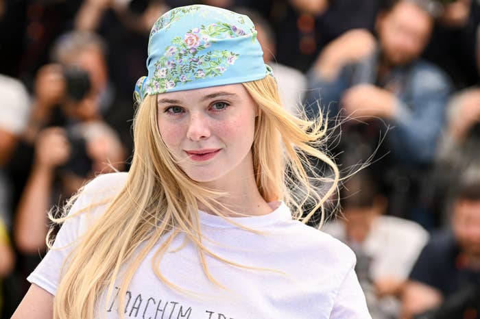Person in a casual white tee and floral headscarf posing confidently at an event, surrounded by photographers on a busy red carpet