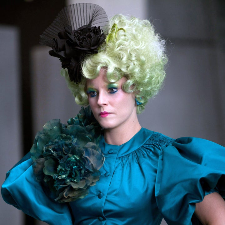 Person in theatrical costume with elaborate ruffled dress and bold accessories, wearing a dramatic hat and styled wig