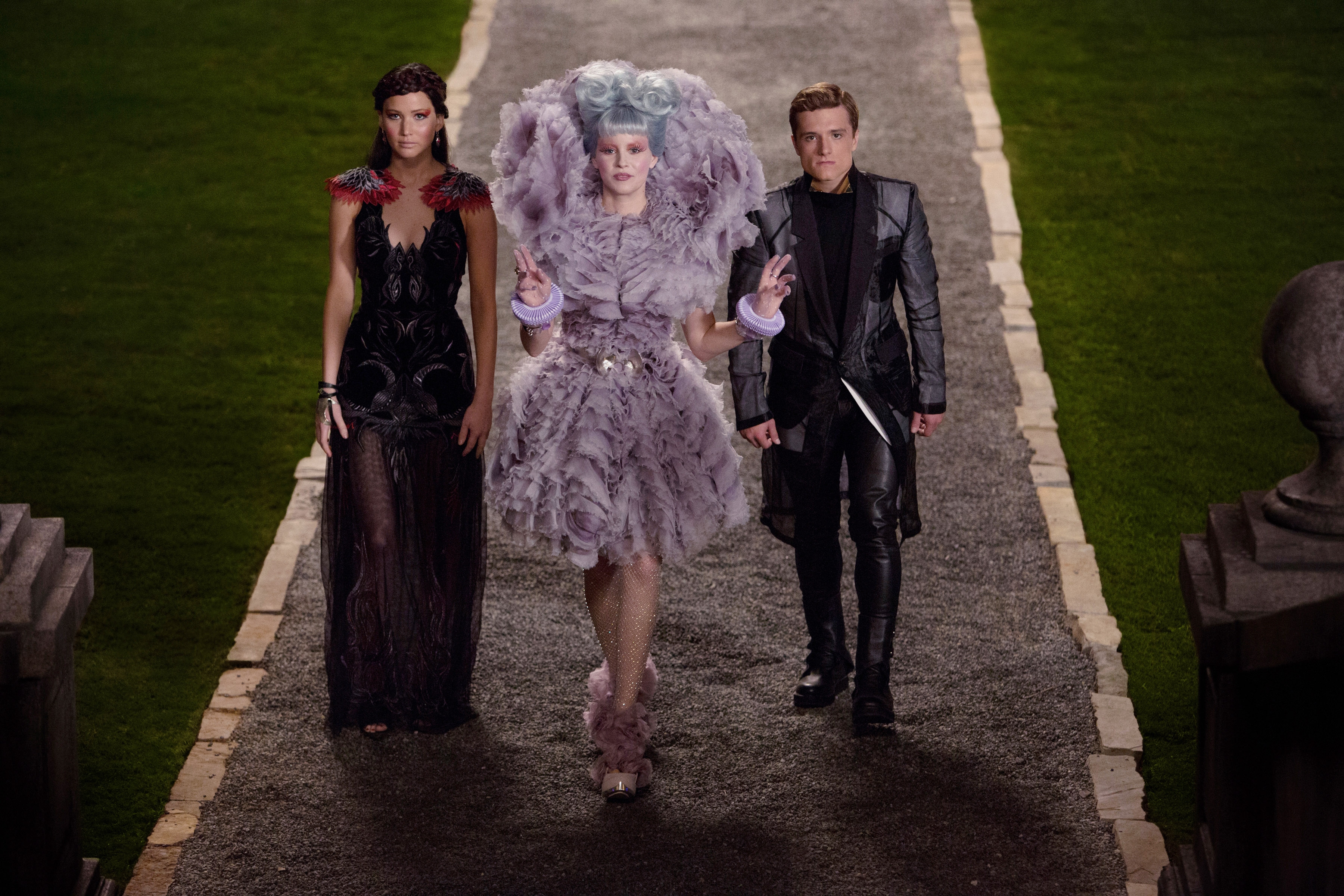 Three people in stylish, extravagant costumes walk on a stone path, evoking a fictional, theatrical setting