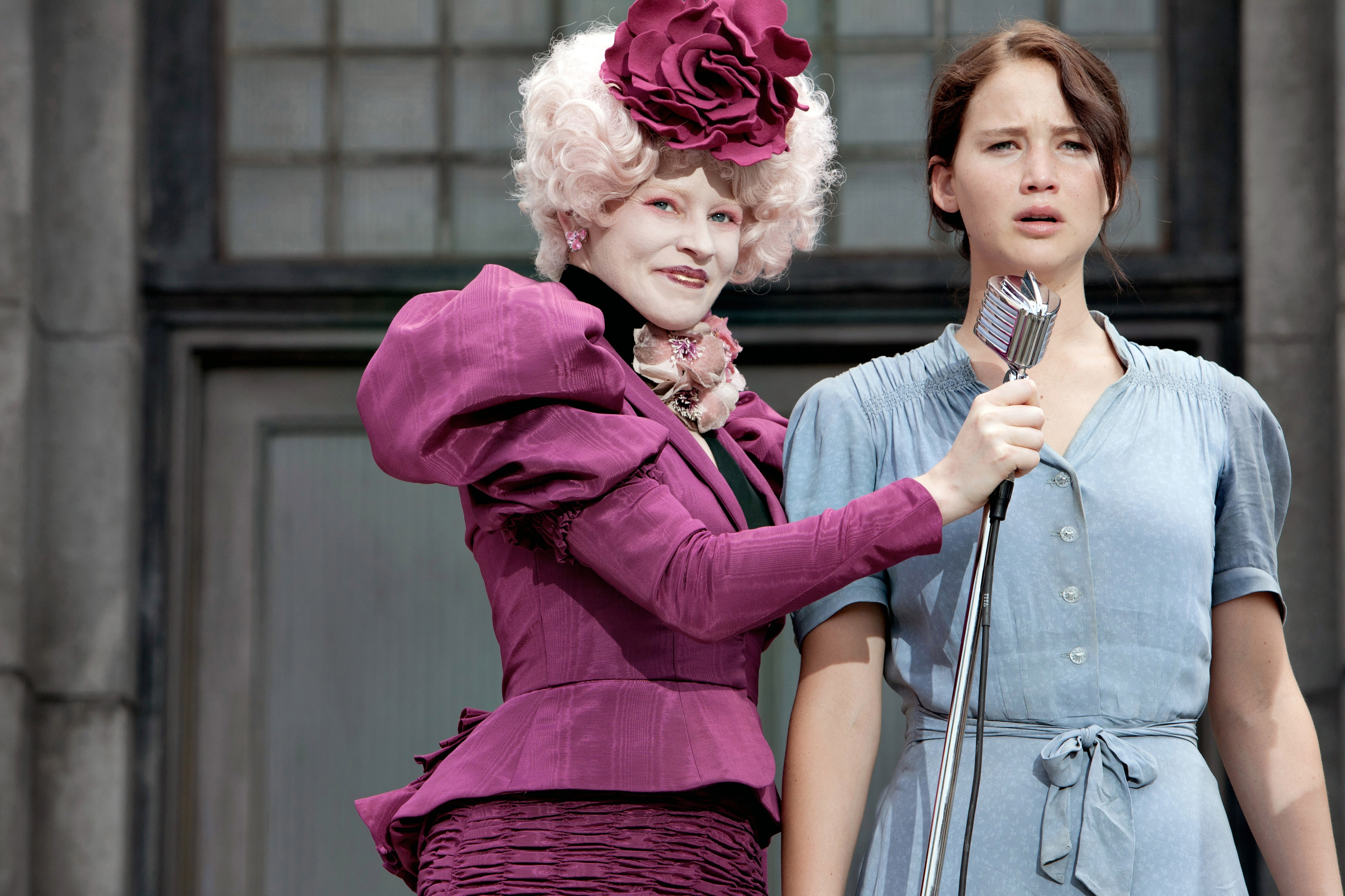 Two characters from a movie scene, one in elaborate period costume and wig with a quirky style, and the other wearing a simple dress, standing with a microphone