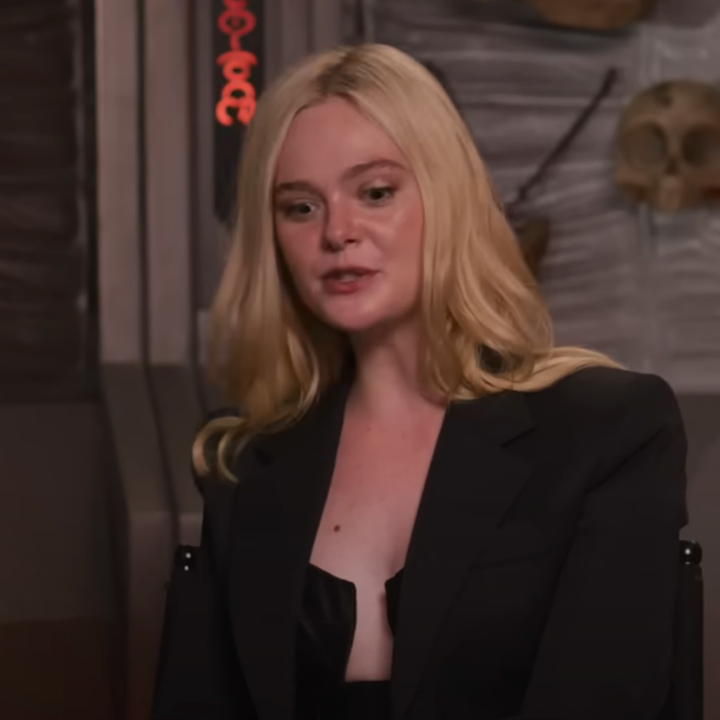 Two people sitting and talking in a studio setting, one in a blazer, with various props like skulls on shelves behind them