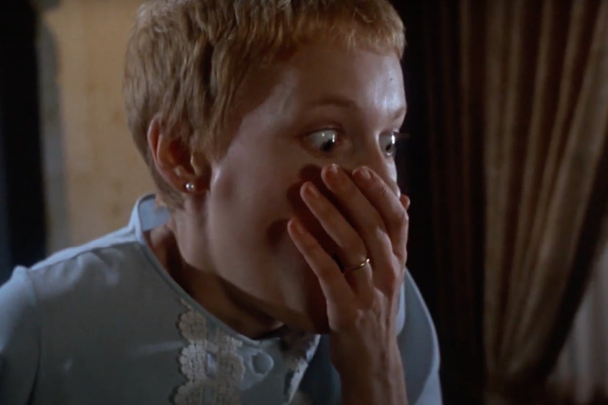 Mia Farrow in "Rosemary's Baby" with short hair, wearing a lace-trimmed top, looks scared with a hand covering her mouth