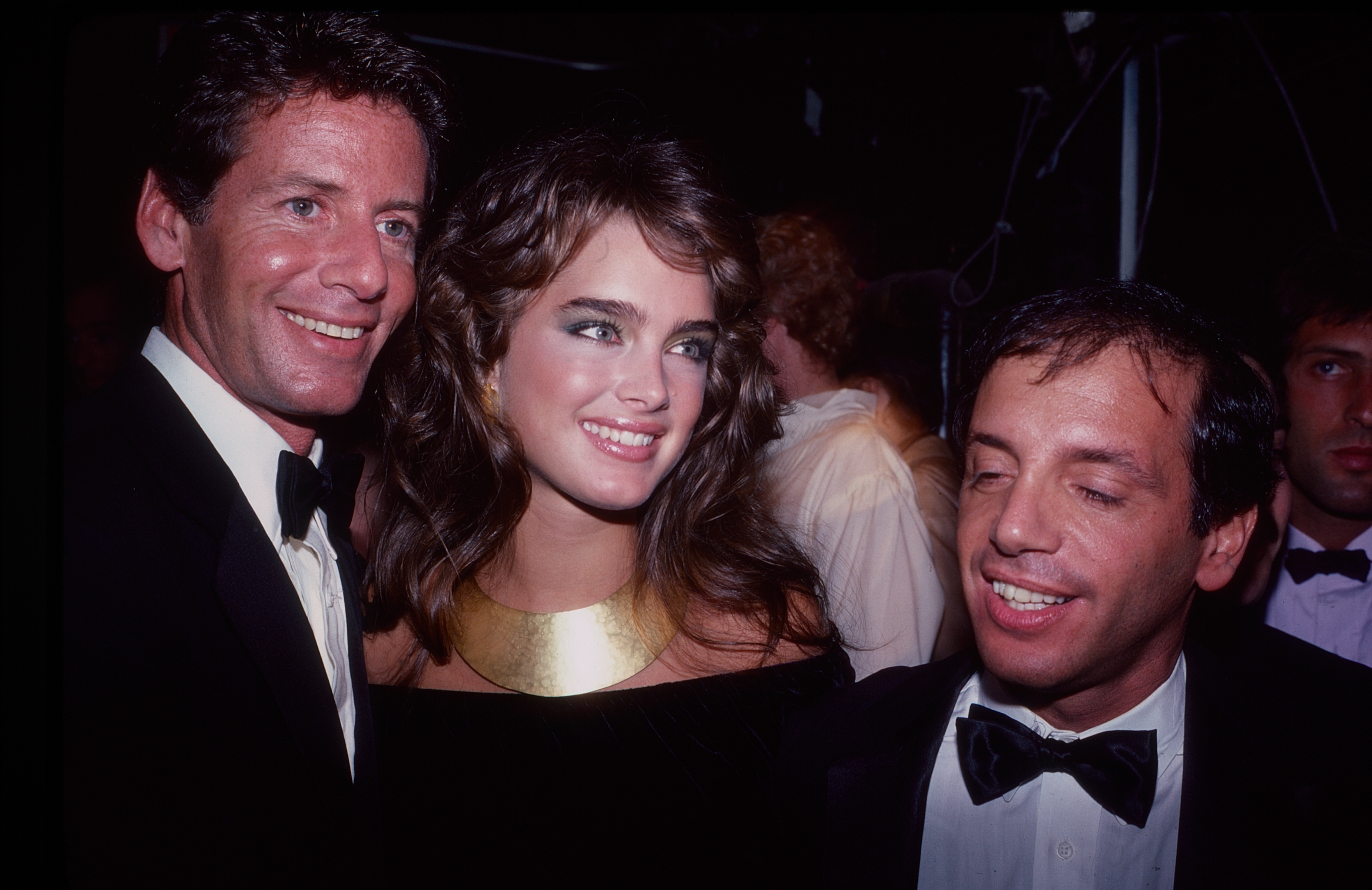 Three people at an event, two in tuxedos and one in an elegant dress with a bold necklace, smiling and posing for the camera