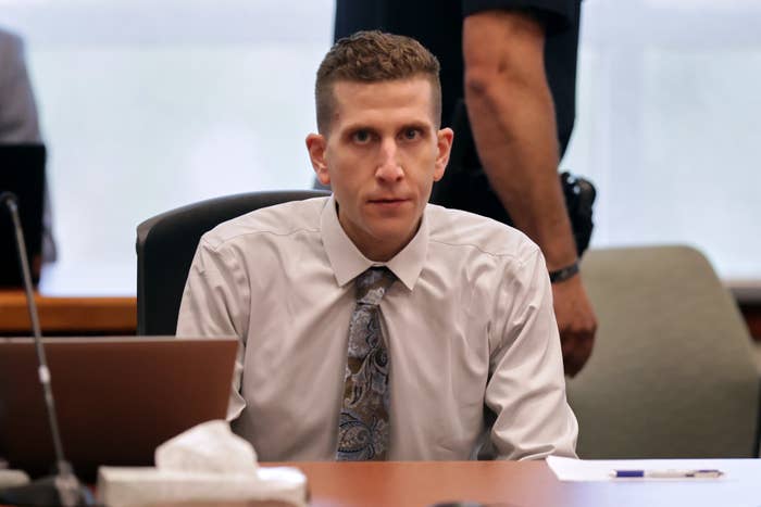 A person in a courtroom sits at a table with a laptop, wearing a patterned tie and light shirt, looking towards the camera