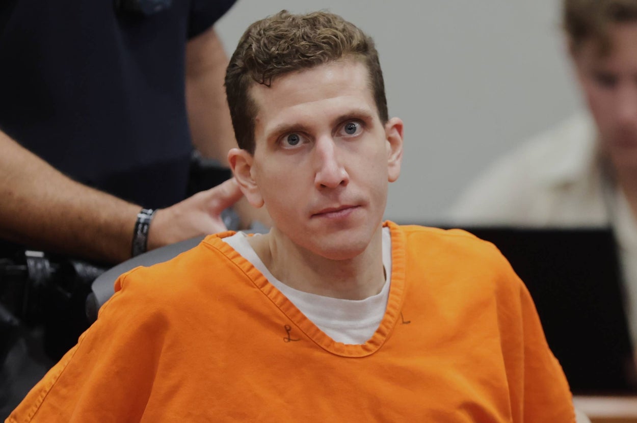 A person in an orange jumpsuit is in a courtroom setting, seated with a serious expression; an officer stands behind them