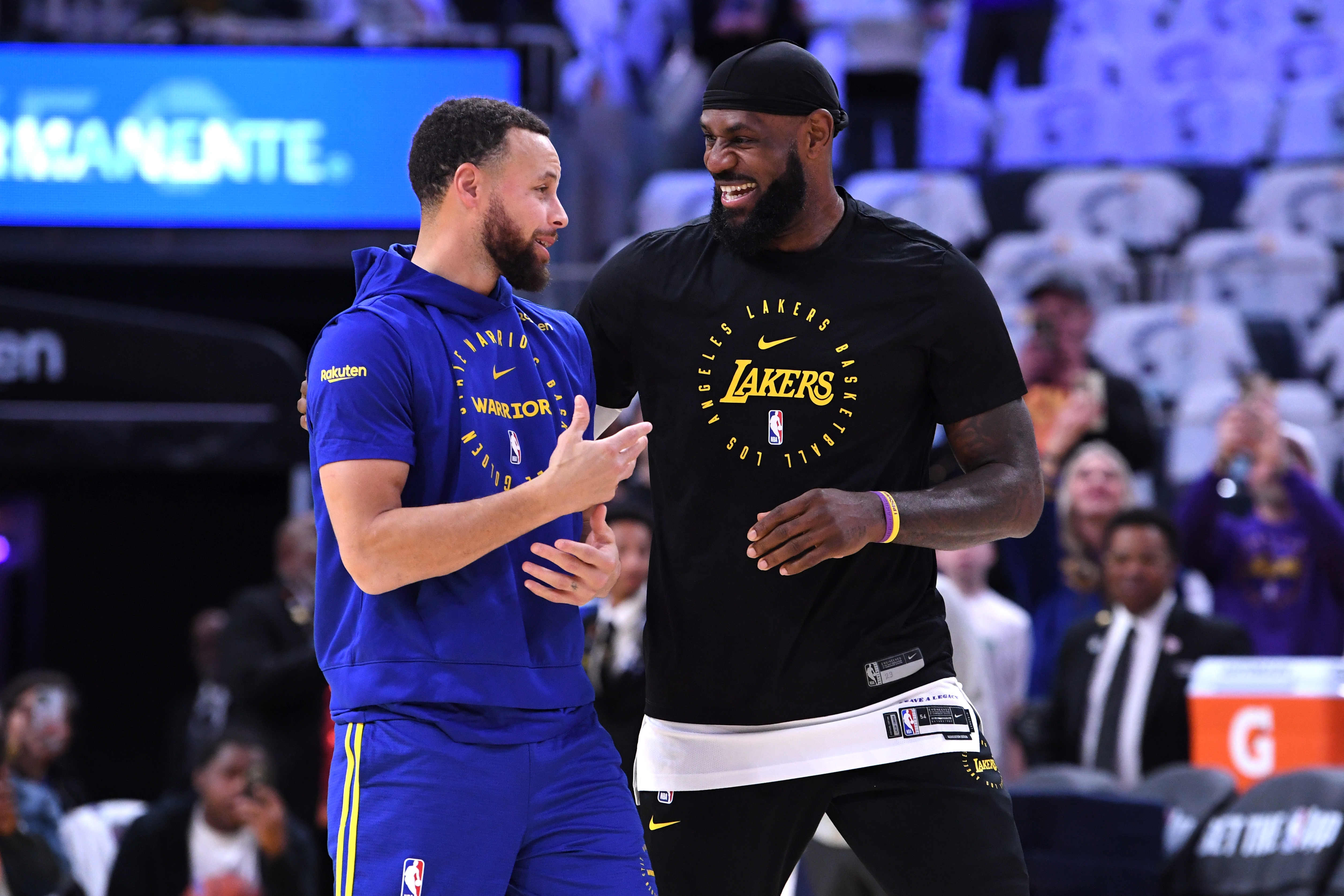 Two basketball players are interacting on the court, one in a blue hoodie and yellow shorts, the other in a black t-shirt and shorts