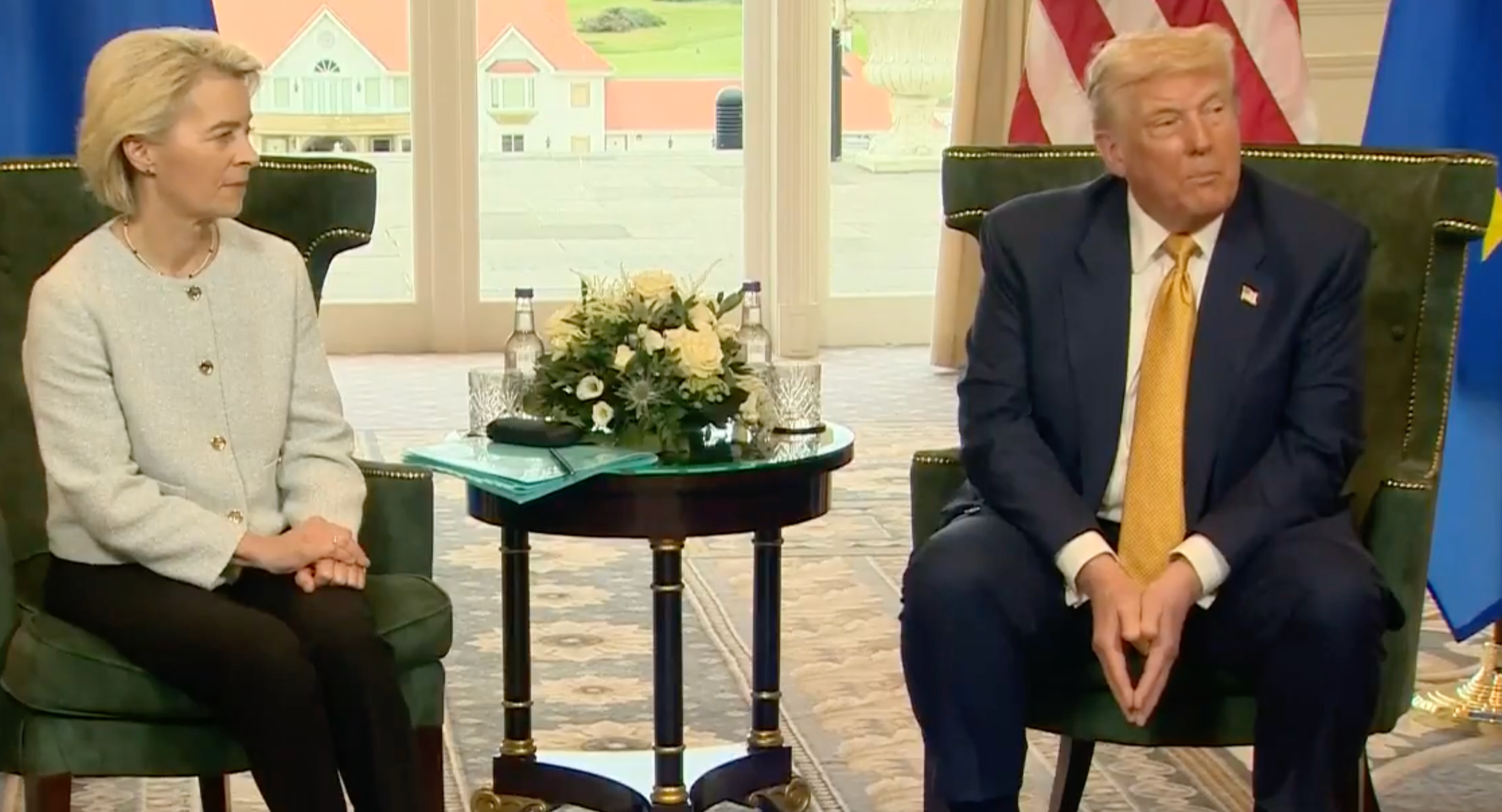 A woman in a formal suit sits on a chair next to a man in a suit with a yellow tie. They&#x27;re in a meeting room with flags in the background