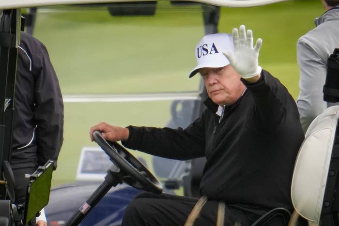 Person in a golf cart, wearing a &quot;USA&quot; cap and raising a gloved hand in a wave, on a golf course