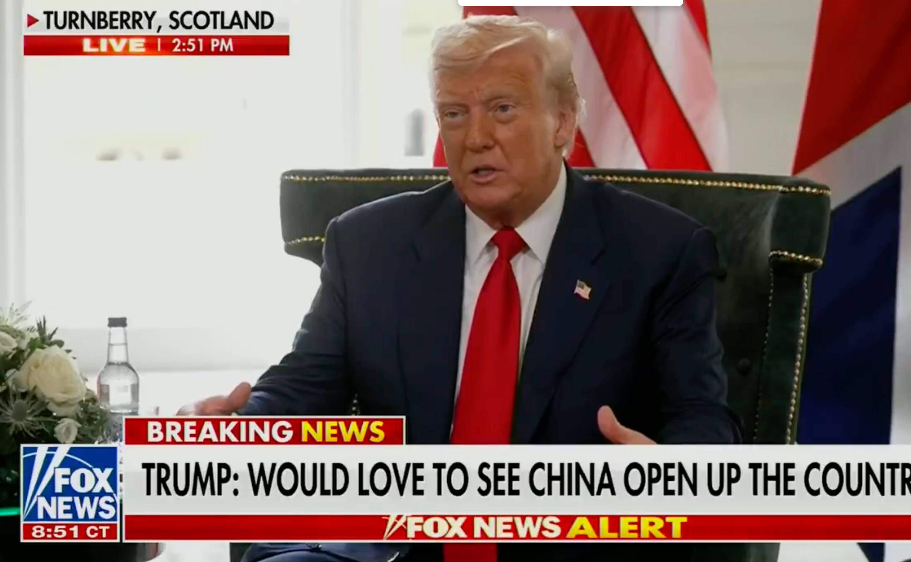 A person is sitting on a chair with flags in the background. The news chyron discusses China and a desire to see the country open up