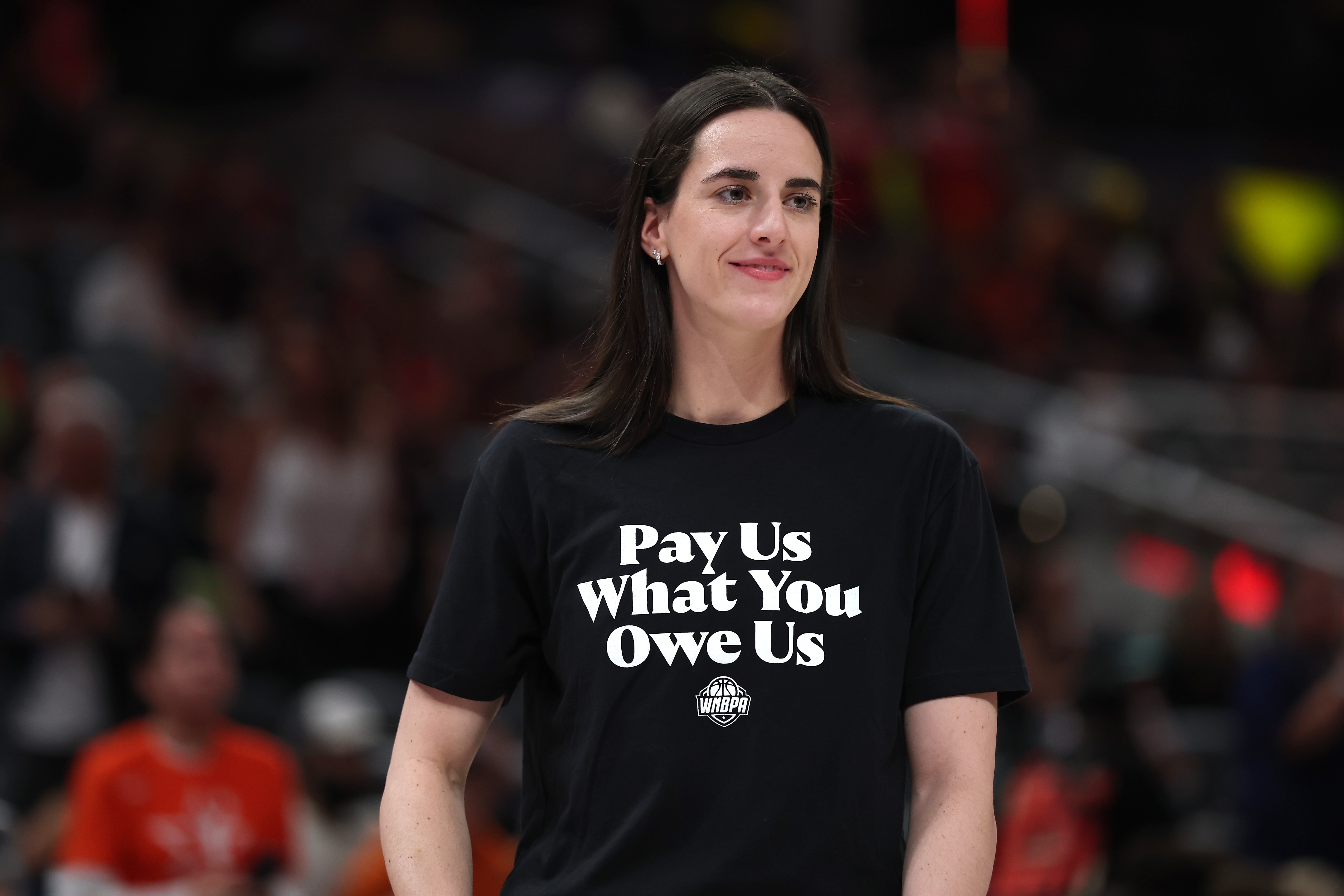 Woman on a stage wearing a t-shirt with "Pay Us What You Owe Us" printed on it, appearing confident and focused