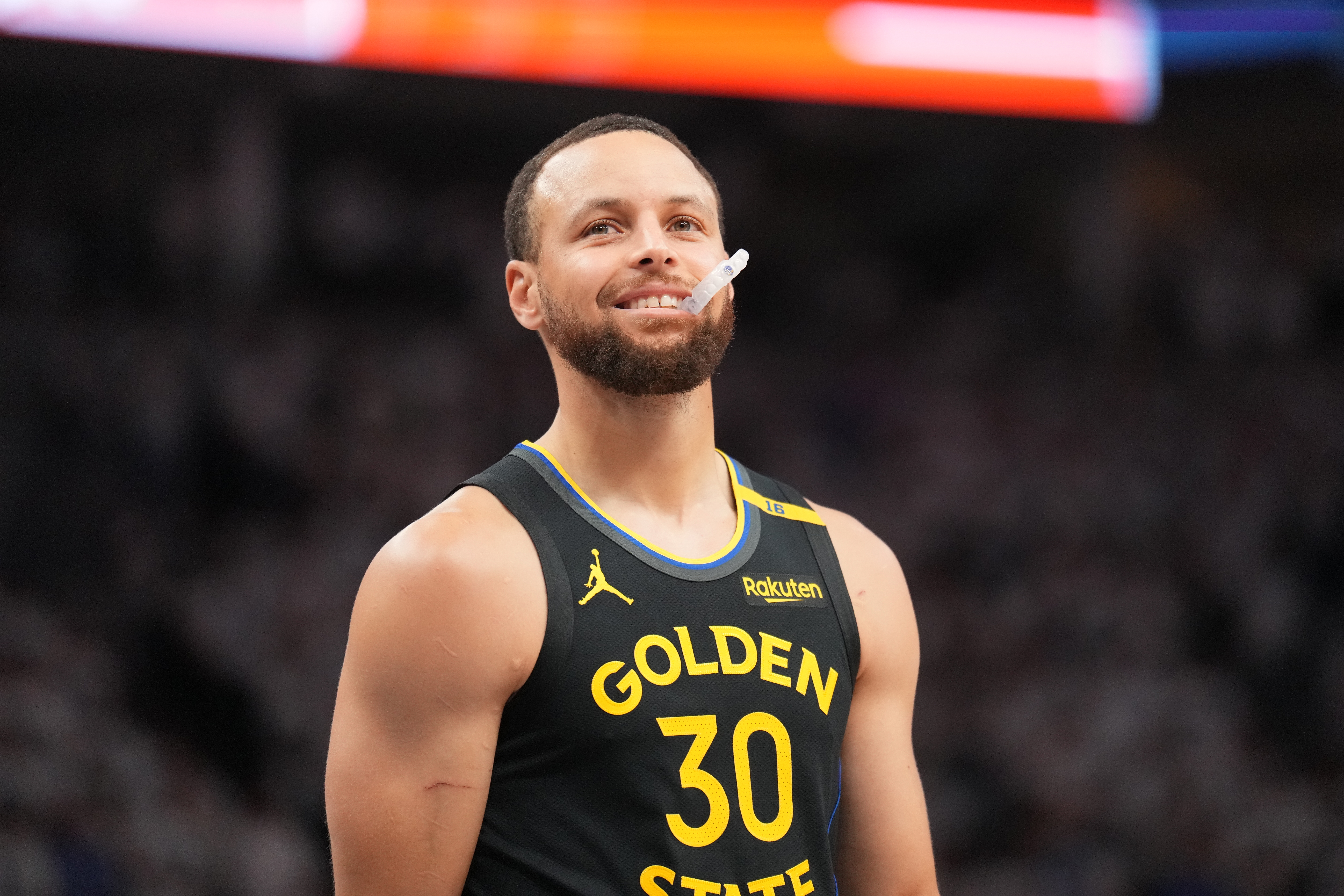 Basketball player in a sleeveless Golden State jersey, holding a mouthguard, standing on the court smiling