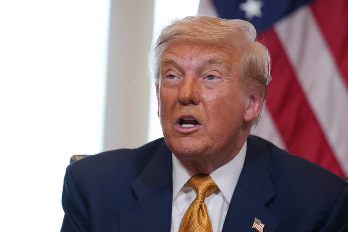 A person in a suit and tie speaks, seated in front of a blurred background with an American flag visible to the side