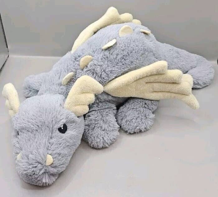 Peluche de dragón gris con alas y detalles blancos, acostado sobre una superficie plana