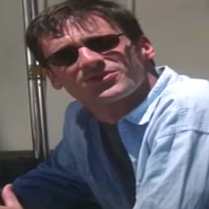 Two individuals sit at an outdoor table wearing sunglasses. One wears a casual black outfit, the other a light shirt, both in a relaxed pose