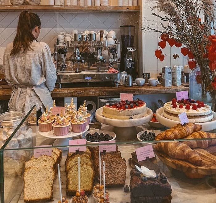 Cafe with desserts like cakes and pastries on display. A person in a casual outfit works at a coffee machine in the background. Cozy decor