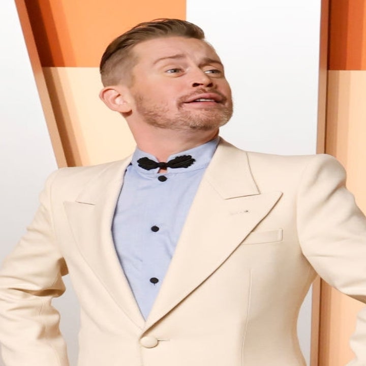 Man in a beige blazer with black bow tie poses confidently on a red carpet backdrop