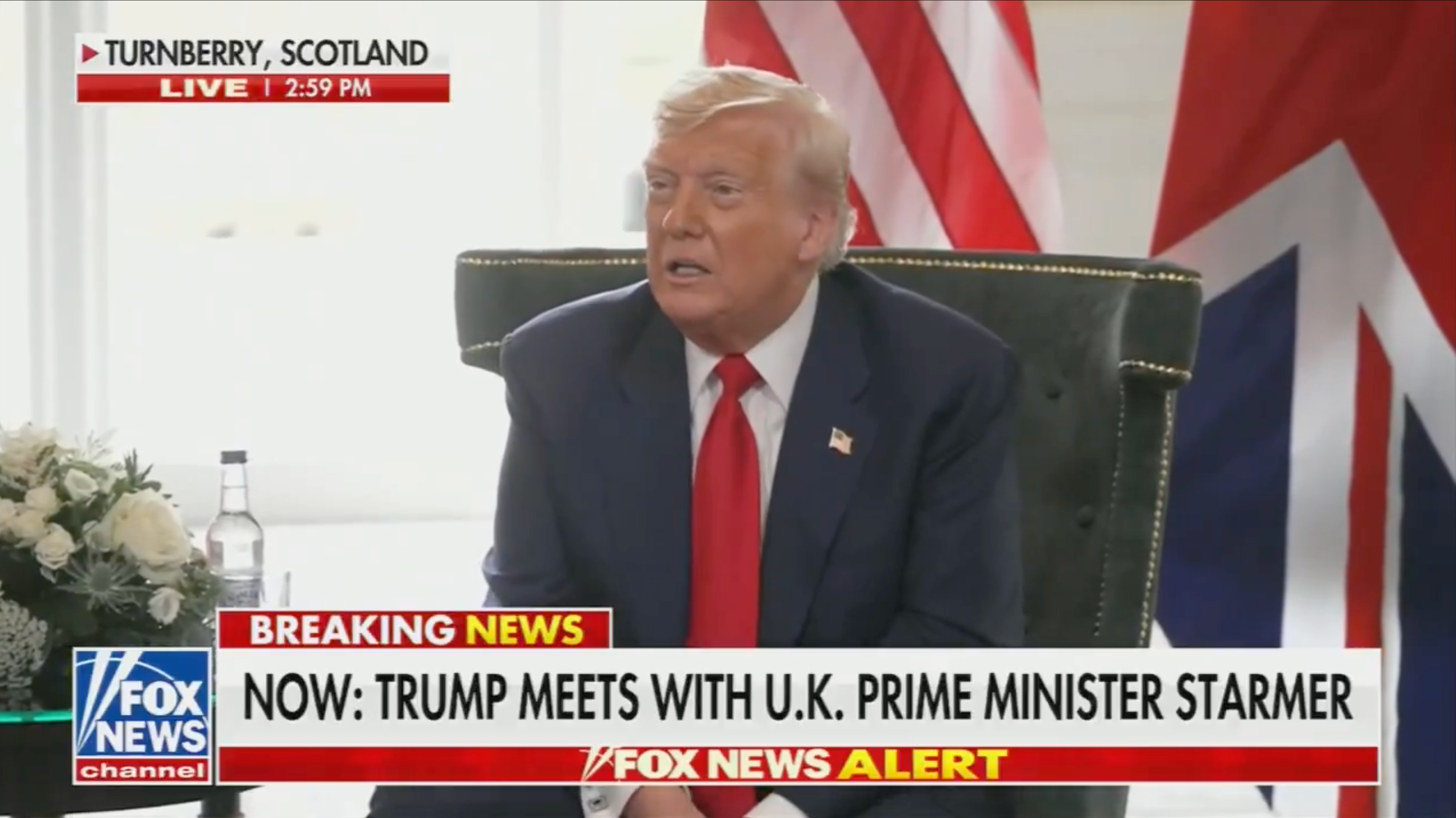 A man in a suit is seated in an interview setting, with a flag in the background. News ticker mentions a meeting with the UK Prime Minister