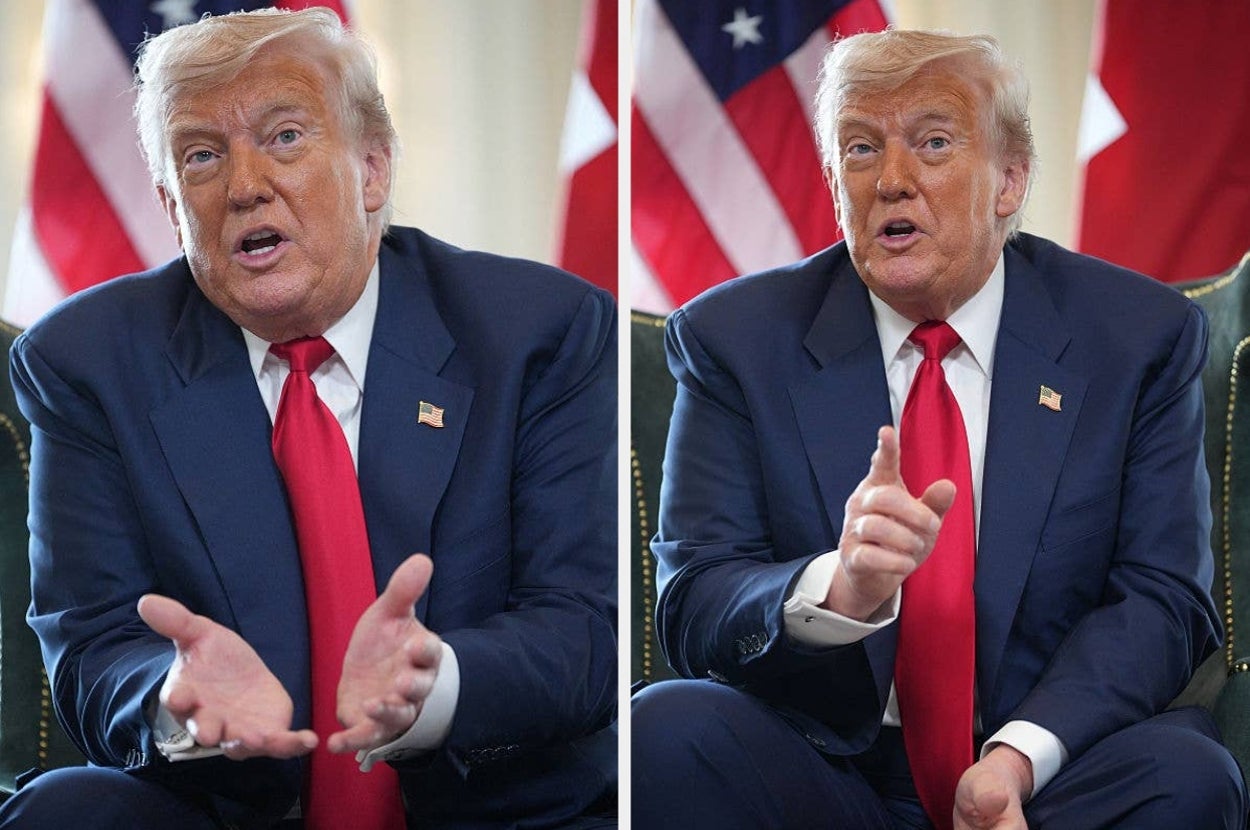 Person in a suit and red tie, gesturing expressively during an interview or discussion, seated indoors with flags in the background
