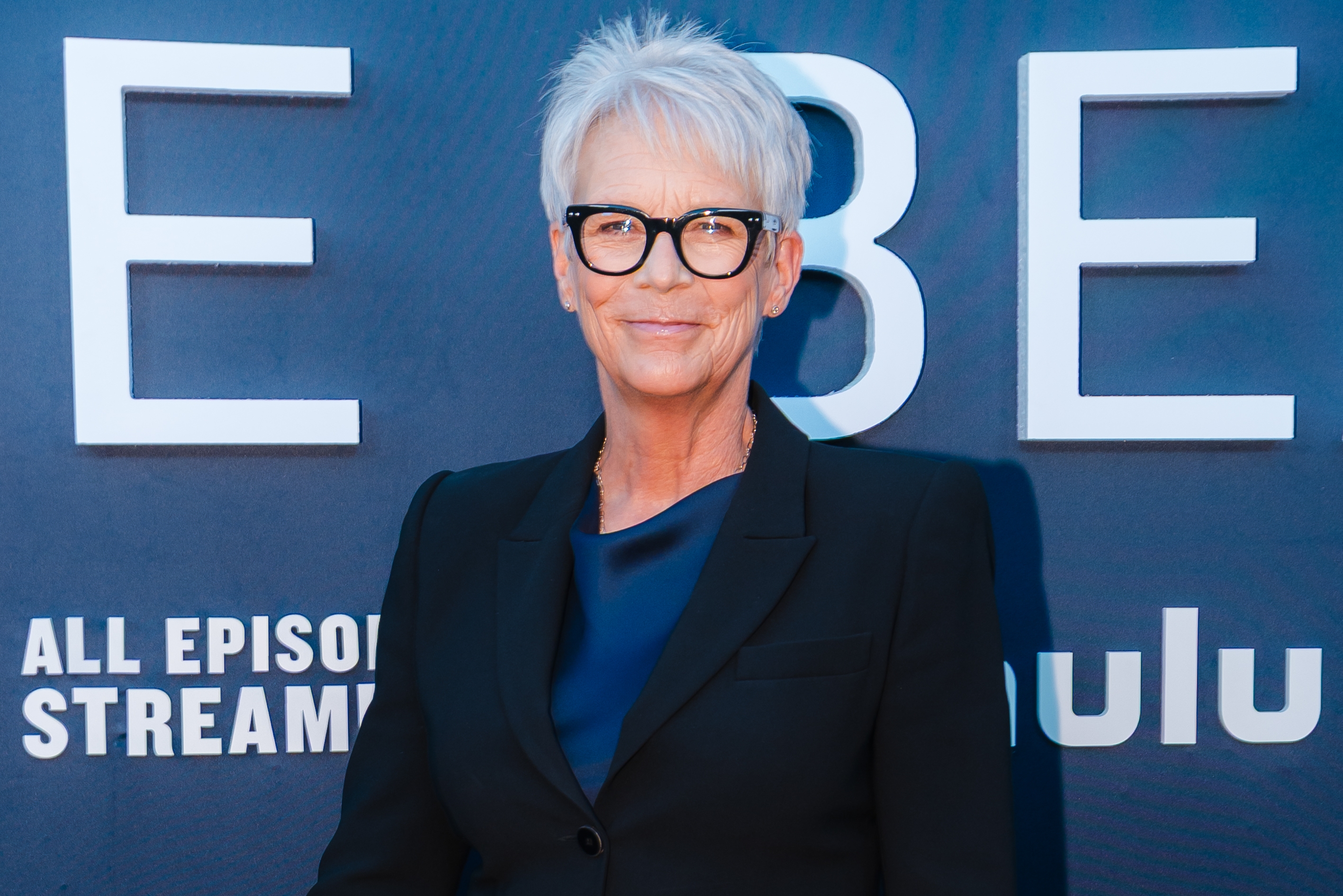Person with short hair in a dark blazer and top, standing on a media event backdrop with text promoting a streaming platform