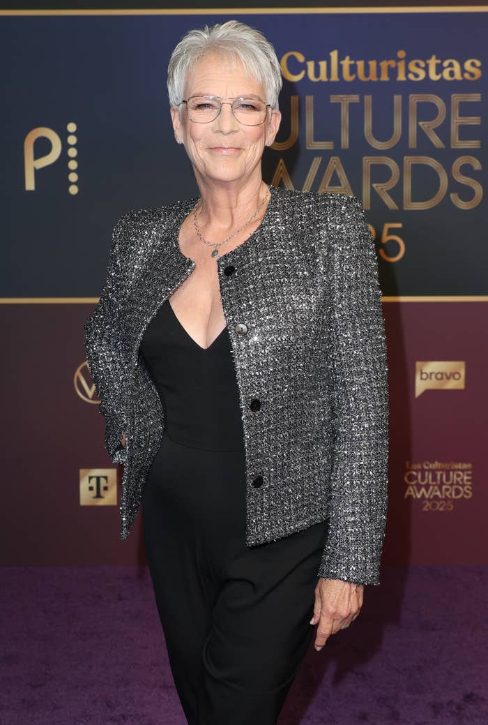 Person in a sparkling jacket and glasses on a red carpet at an event, smiling confidently. Event signage displayed behind them