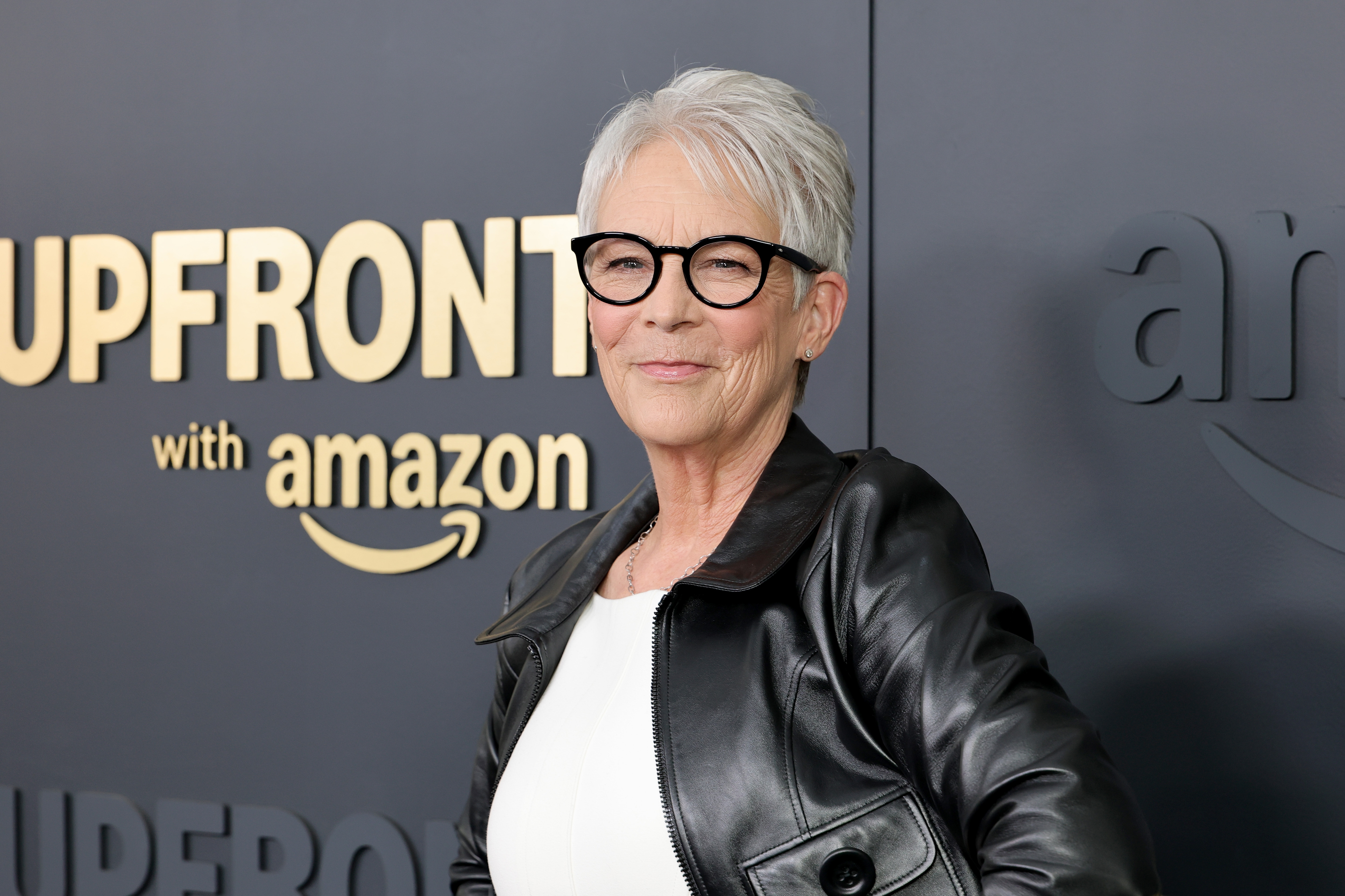 Person in a leather jacket and glasses poses confidently at an event backdrop with &quot;Upfront&quot; and &quot;Amazon&quot; logos