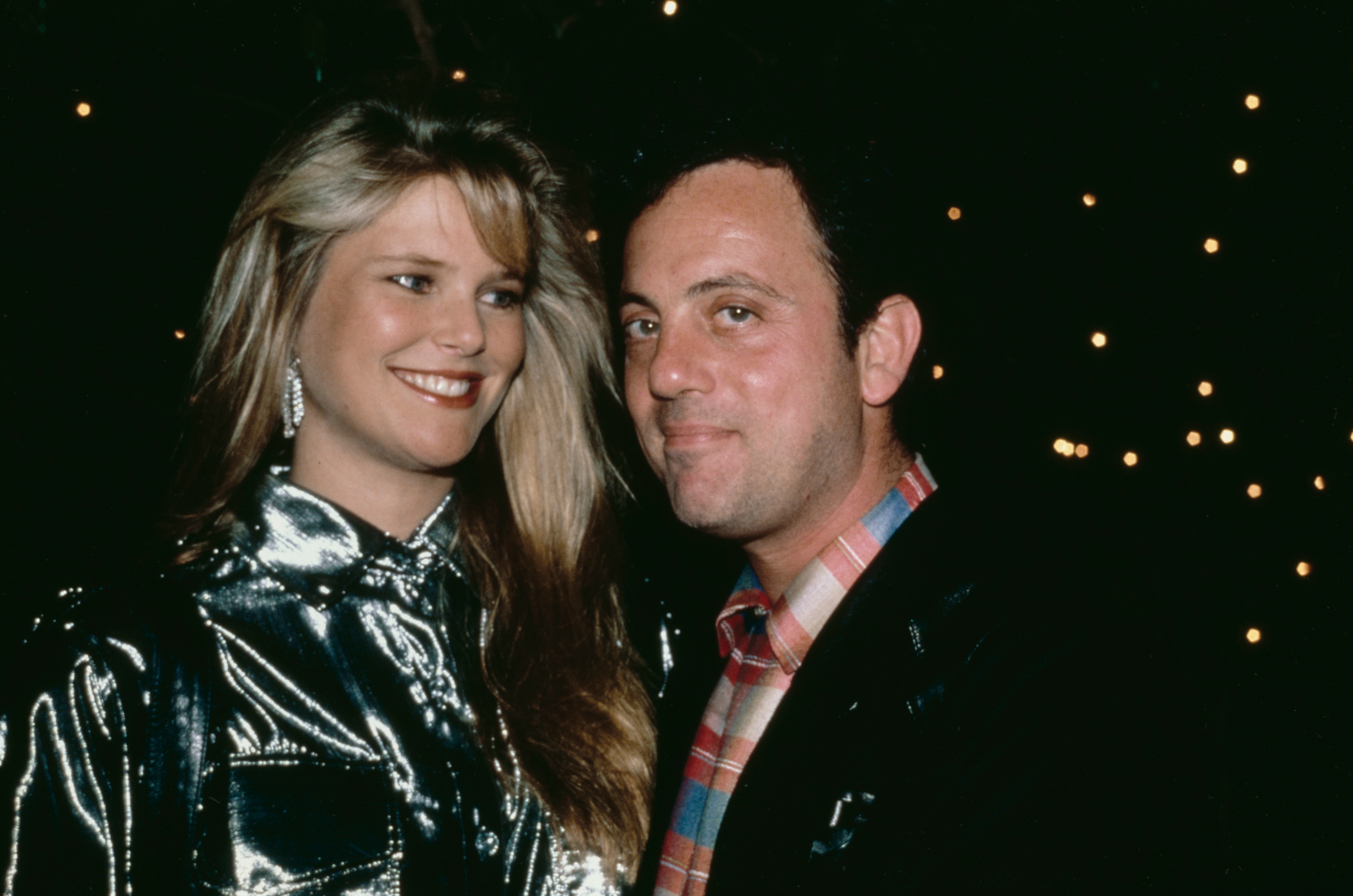 A man and woman at a nighttime event. The woman is wearing a shiny, metallic top, and the man is in a plaid shirt with a dark jacket