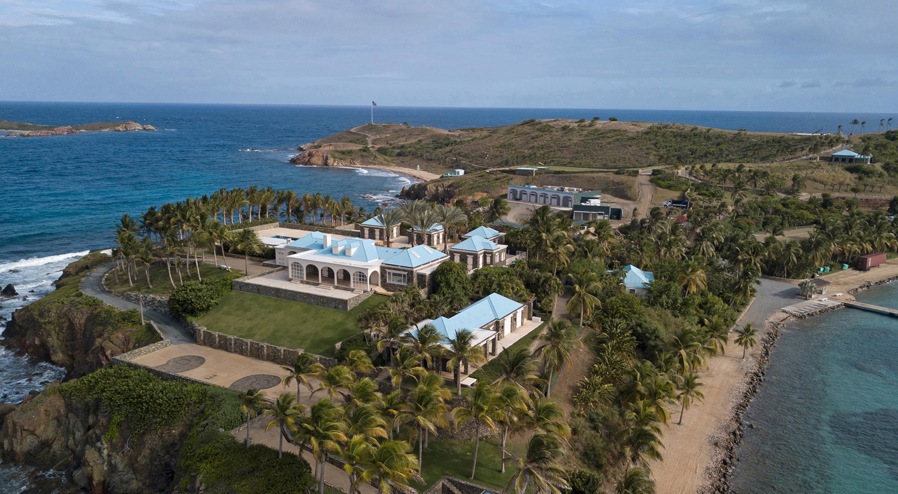 Aerial view of an island estate surrounded by palm trees, with ocean waves hitting the shore and multiple buildings visible on the property