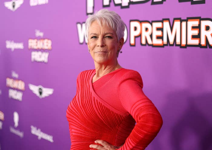 Person posing confidently on a red carpet, wearing an elegant, form-fitting gown with unique shoulder and neckline details