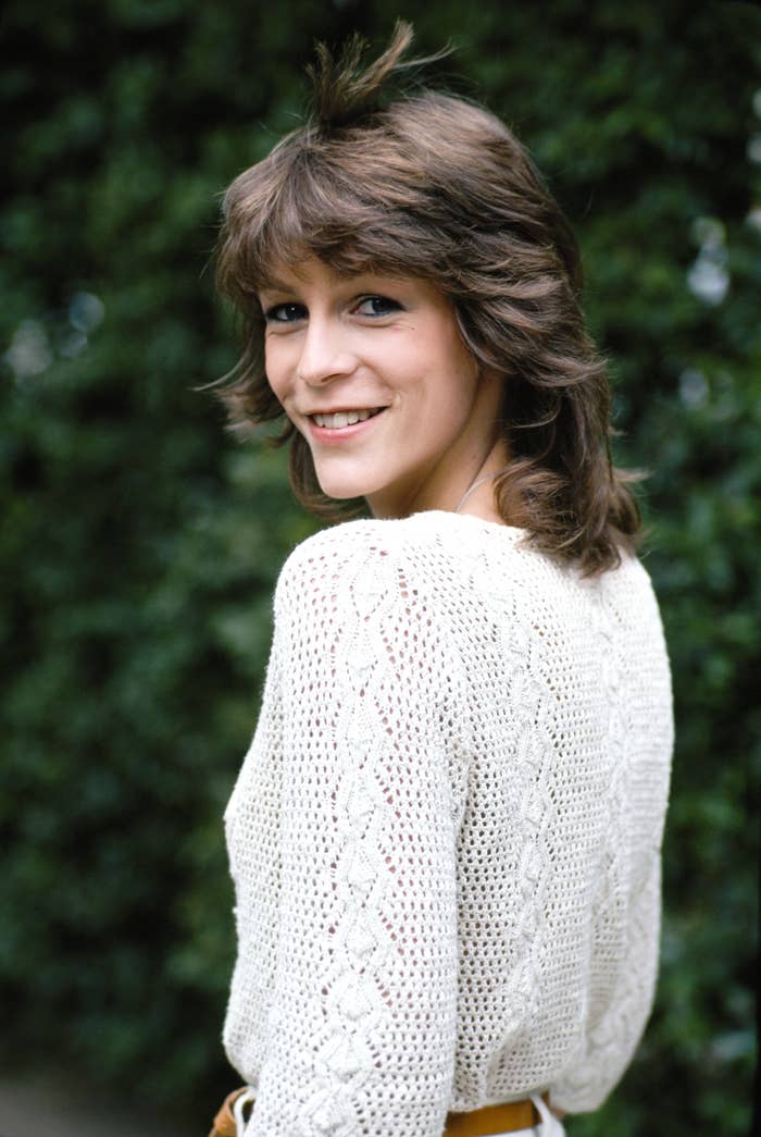 Person with layered hairstyle smiling, wearing a white knit sweater, looking over shoulder outdoors