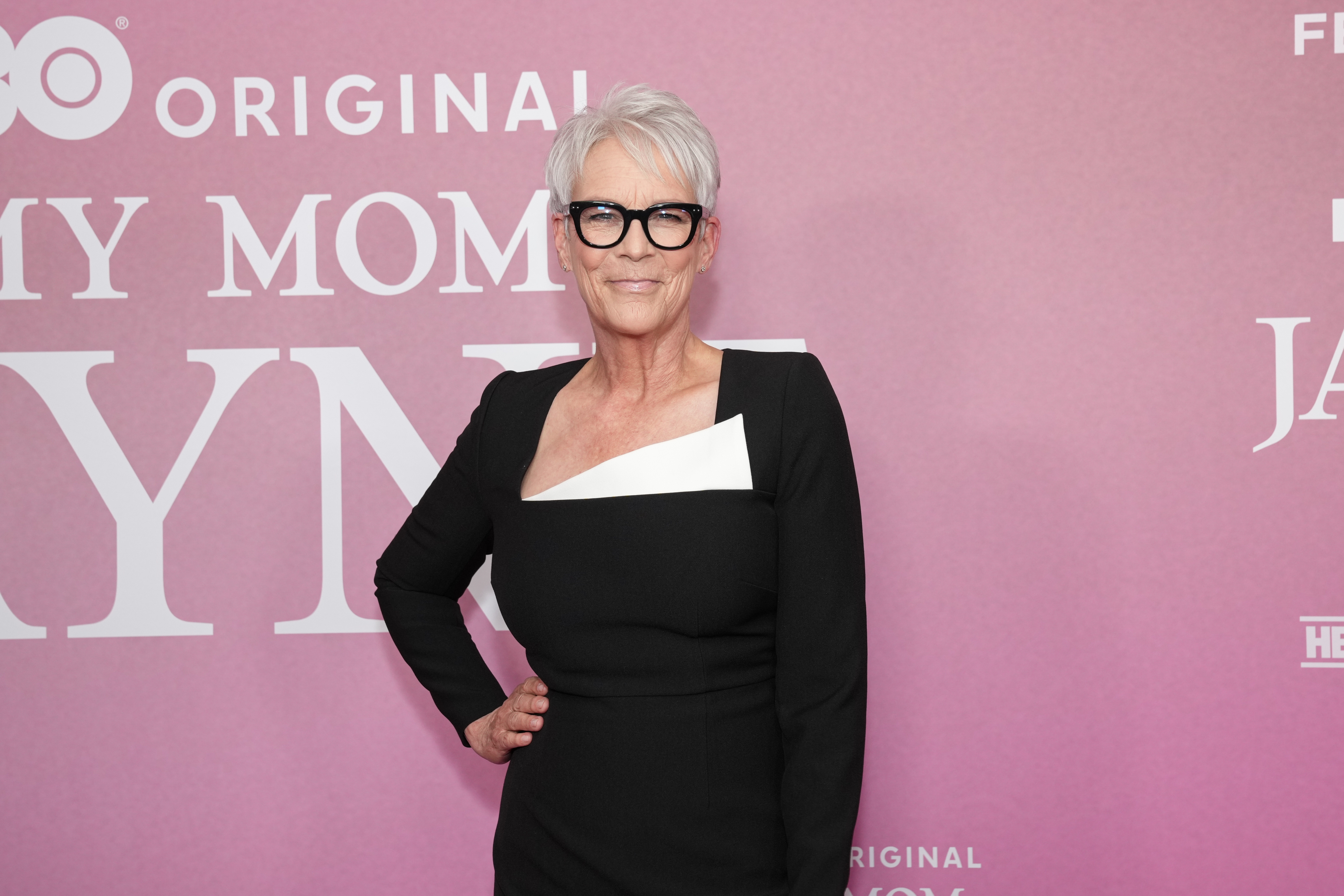 Person in an elegant, black and white dress at a red carpet event, wearing glasses, against a pink backdrop with text partially visible