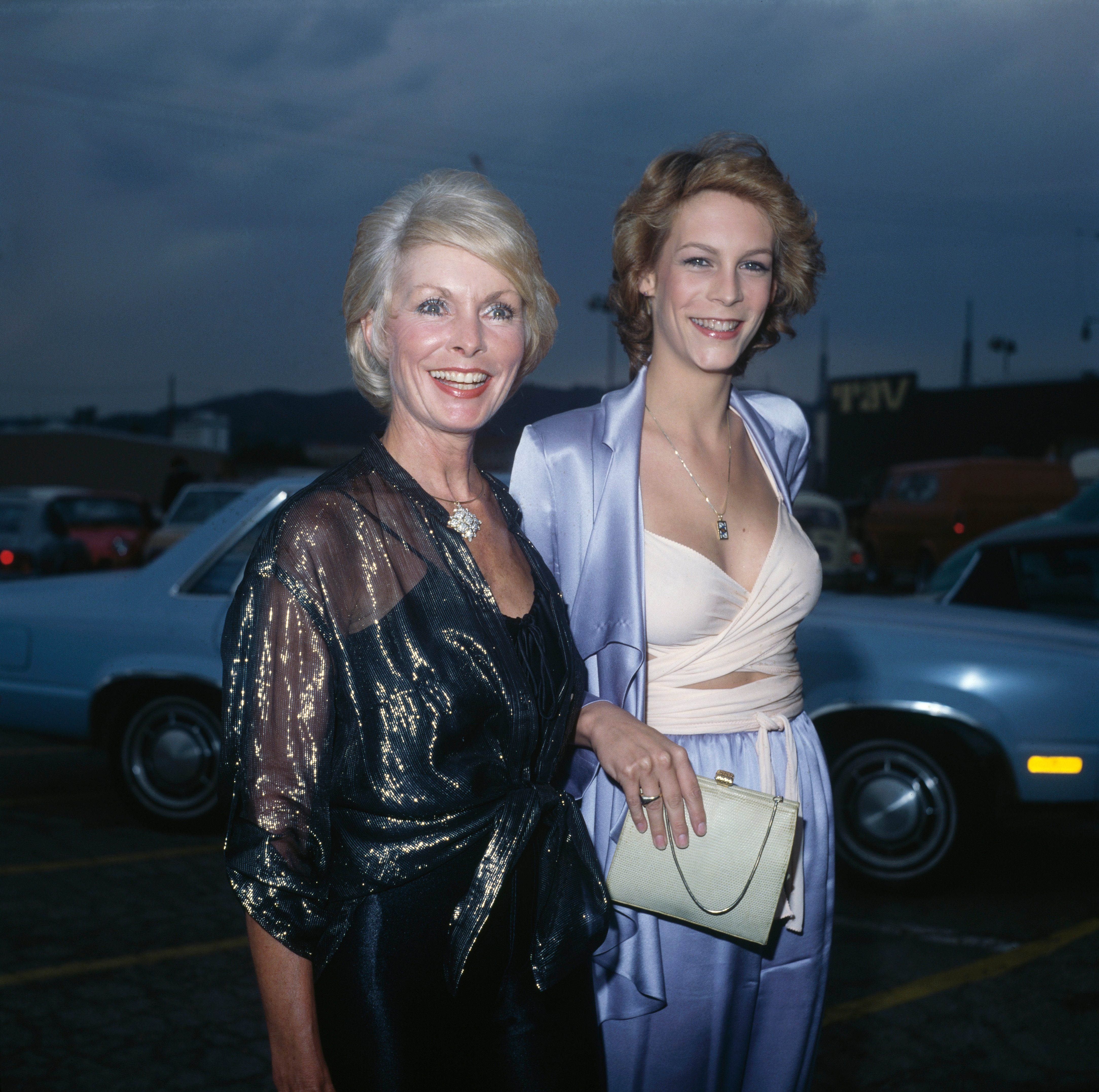 Two people smiling at an outdoor event. One is wearing a shimmering dark outfit, the other in a light suit with a draped top, holding a small clutch
